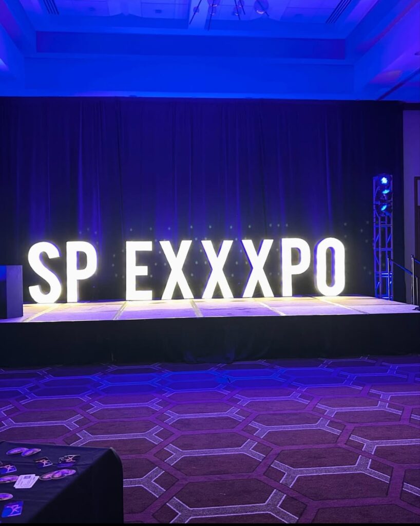 Illuminated "SP EXXXPO" marquee letters on stage at the Southern Pleasure Expo, set against a dark curtain with blue uplighting in a hotel ballroom setting.